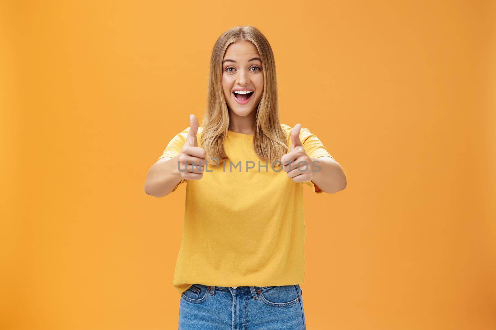 Woman supports with raised thumbs up and amused cheerful smile showing positive attitude expressing like on concept or idea giving approval posing happy and delighted against orange background by Benzoix