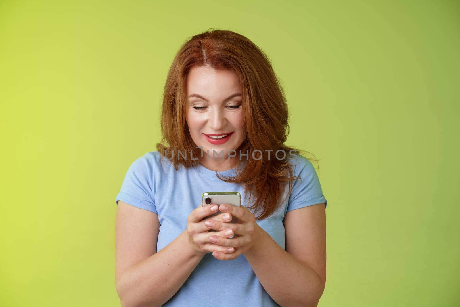 Intrigued excited tempting redhead middle-aged woman shopping online smartphone hold mobile phone look pleased entertained gadget screen smiling delighted playing game found perfect app.