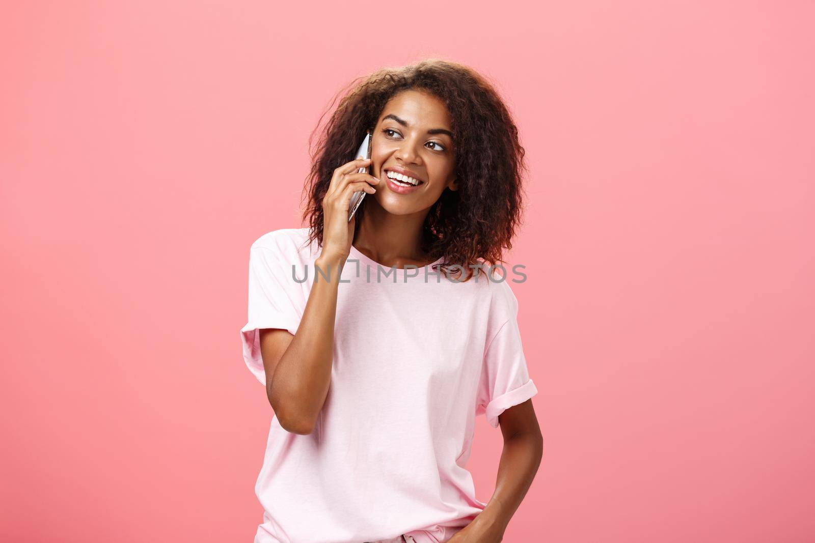 Girl calling friend to meet up. Portrait of charming friendly and outgoing african american young woman with afro hairstyle holding smartphone near ear while talking looking left curiously. Copy space