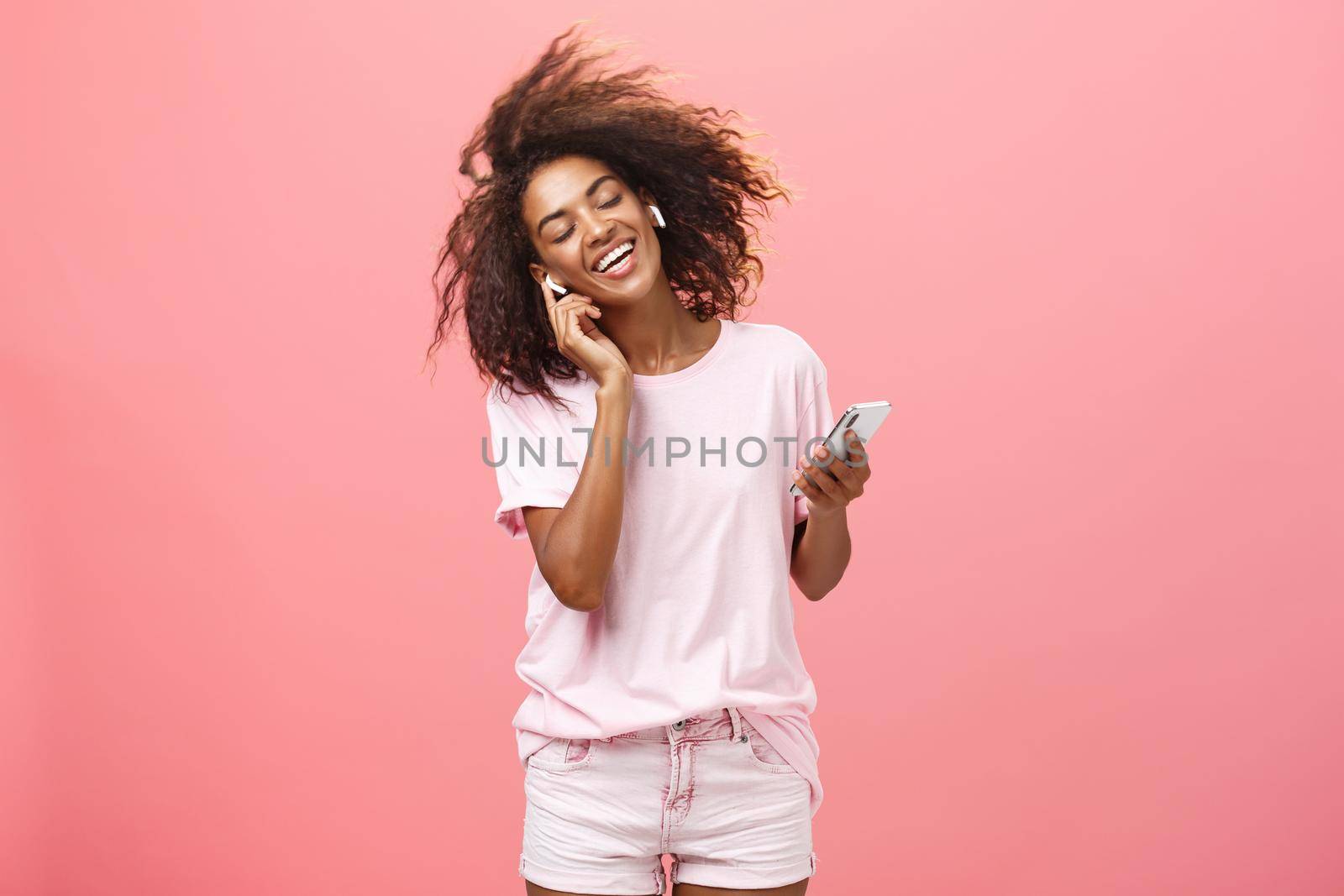 Getting rid of worried with good tunes in ears. Pleased attractive young african woman in trendy outfit listening music in wireless earphones touching earbud smiling dancing and holding smartphone by Benzoix