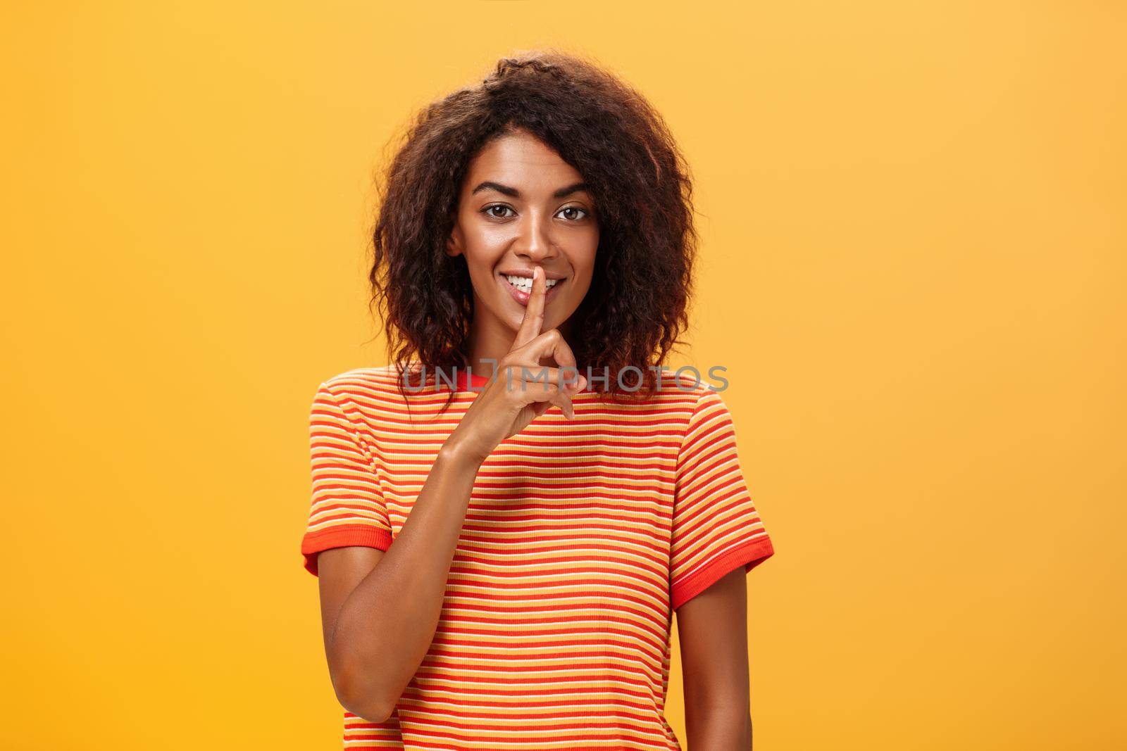 Not say anyone till I give permission. Happy pleased and mysterious attractive dark-skinned female with curly hairstyle telling beauty secret to friend making shh sound with index finger over mouth.