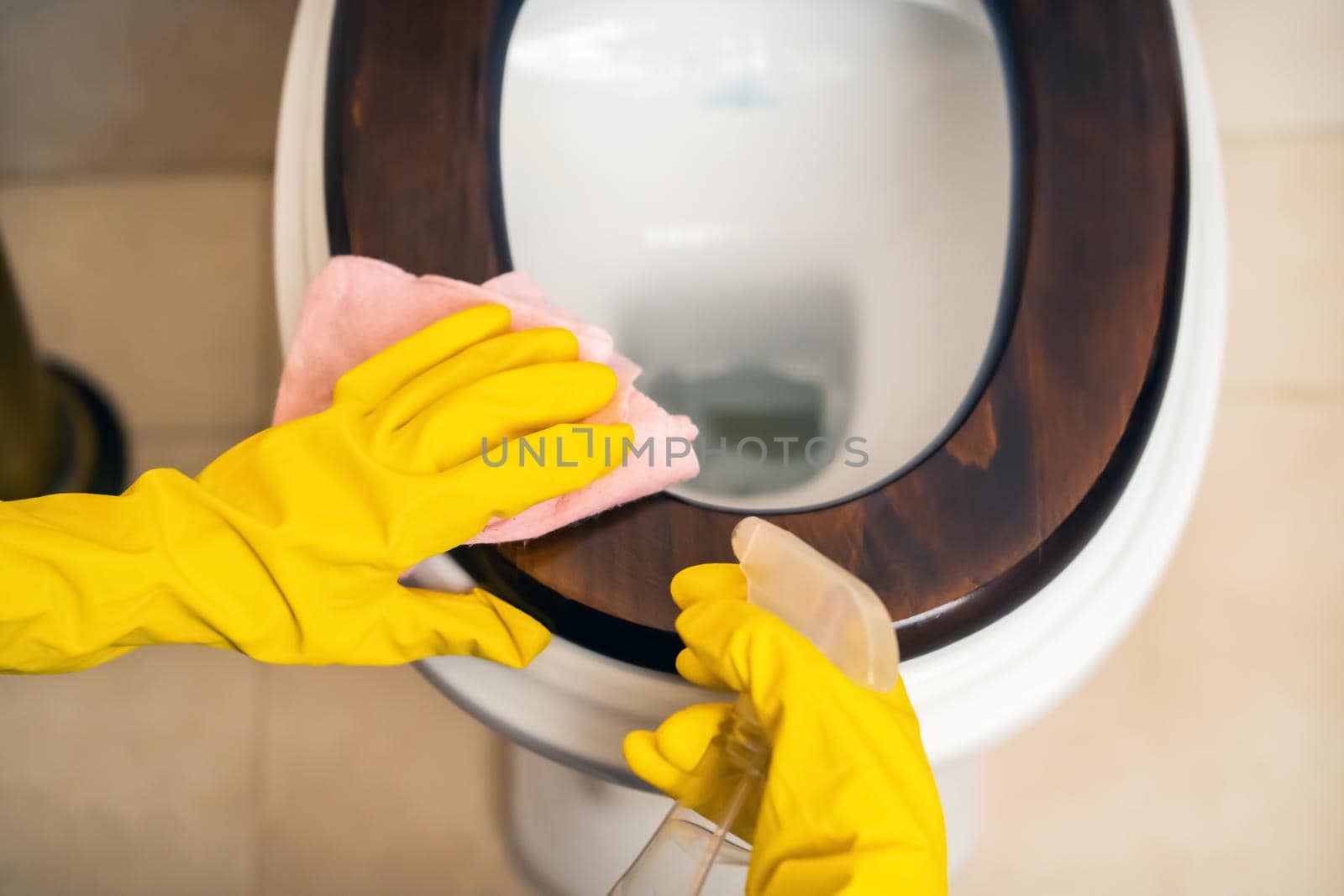 Female hands in yellow rubber gloves are holding a rag and wiping the toilet with detergent and disinfectant. The woman is cleaning the bathroom.