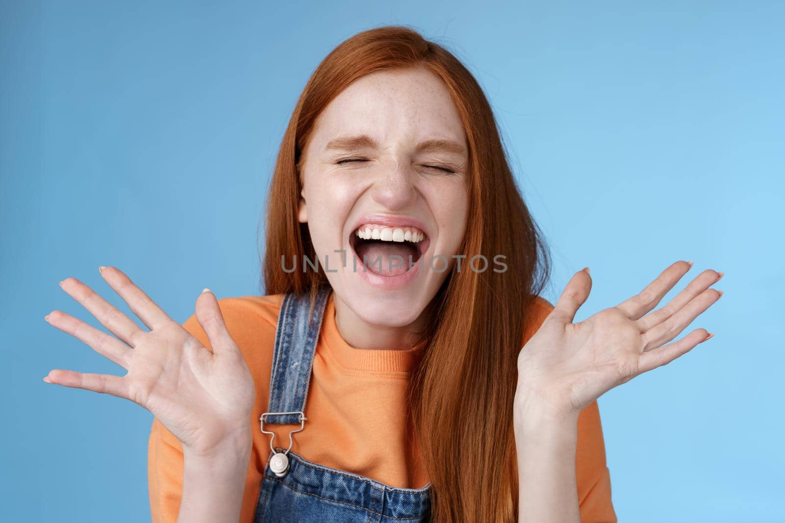 Positive carefree triumphing young attractive redhead girl scream out loud joy happiness close eyes waving raised hands win first prize dream come true celebrating victory good news, blue background by Benzoix