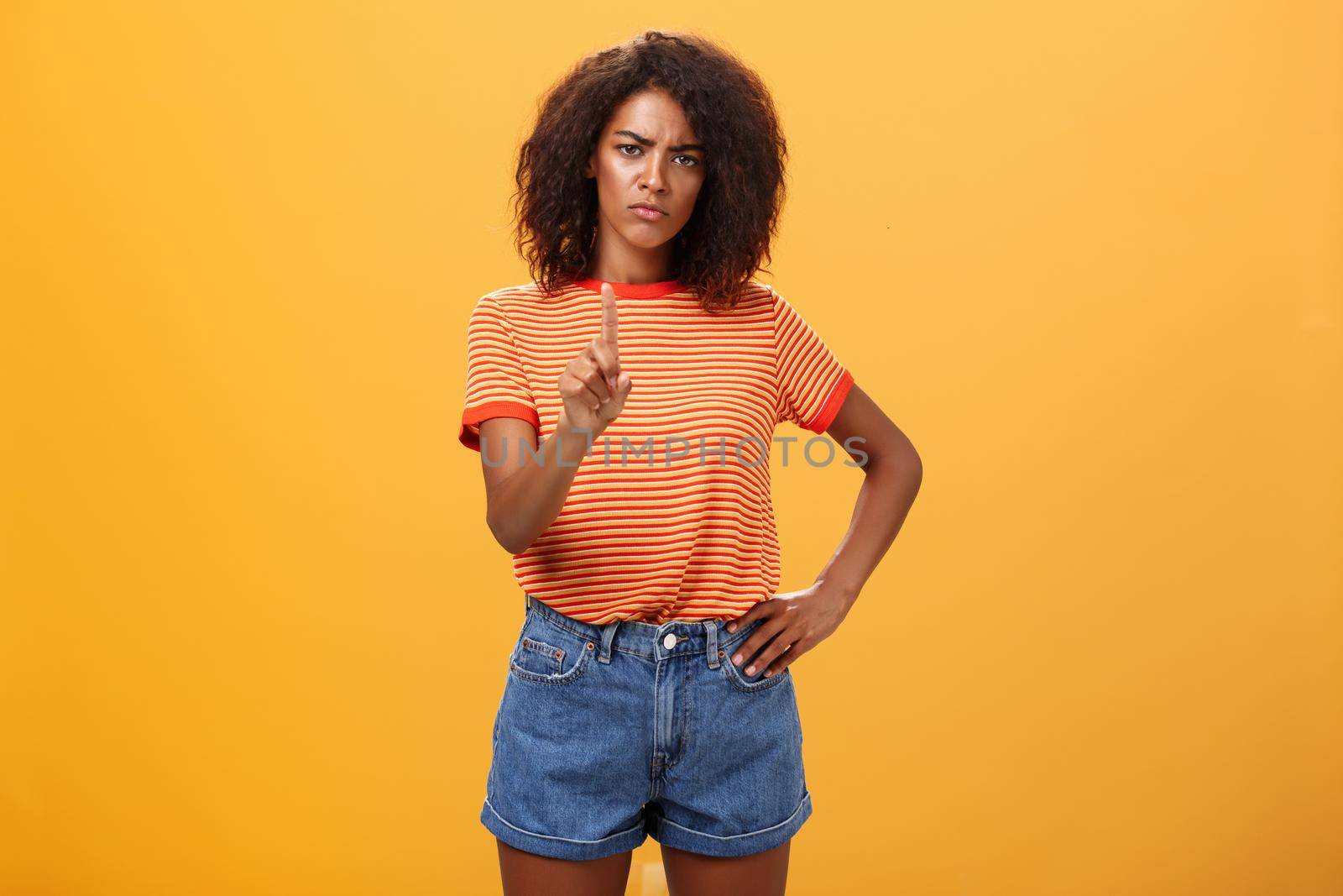 You better not. Dissatisfied bossy serious-looking african american curly-haired female head manager in t-shirt frowning shaking index finger in forbid or stop gesture scolding dog over orange wall. Body language concept