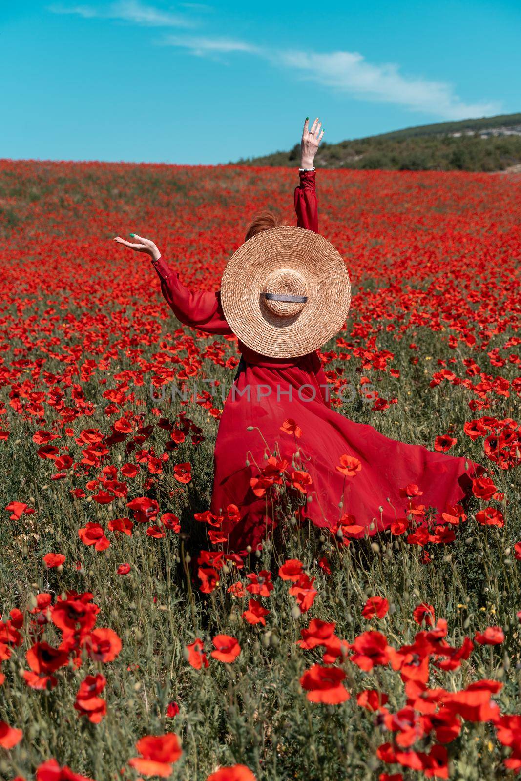 Young woman stands with her back in a long red dress and hat, posing on a large field of red poppies by Matiunina