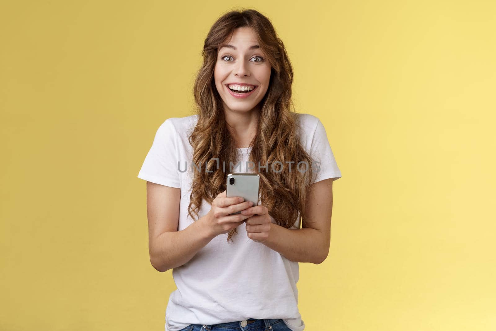 Extremely happy enthusiastic surprised joyful lovely feminine girl receive pleasant touching message online hold smartphone smiling broadly look camera joyful amused yellow background.