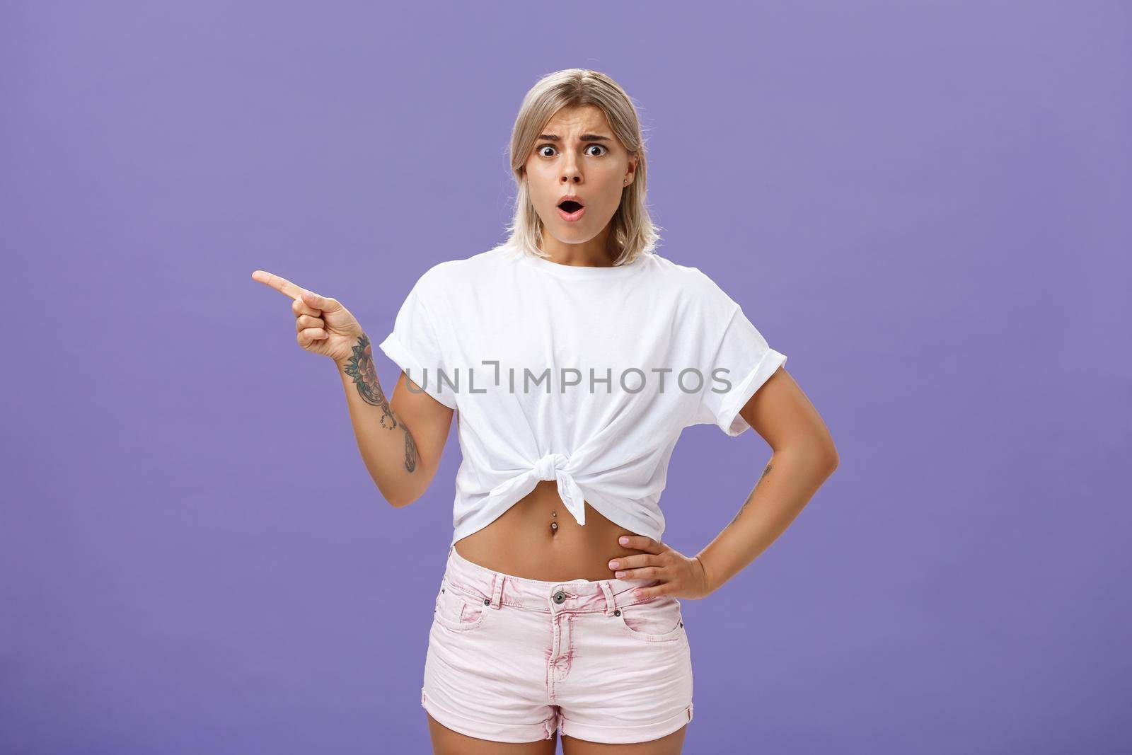 Girl being unhappy with unfair thing happened. Shocked displeased complaining, confused woman with tanned skin blond hair and tattoos opening mouth and frowning from jealousy or regret pointing left.