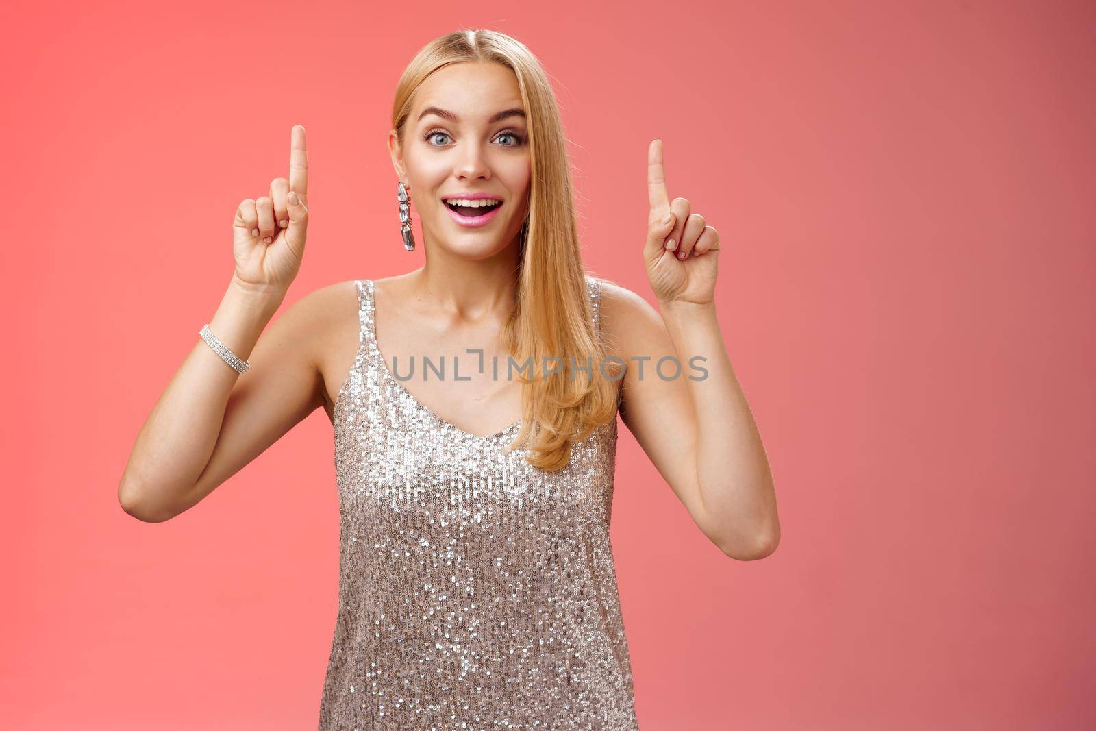 Charming amazed blond european woman in fabulous silver glittering dress raise hands point up amused enjoying watching shooting stars, fireworks gaze camera excited happy surprised, red background.