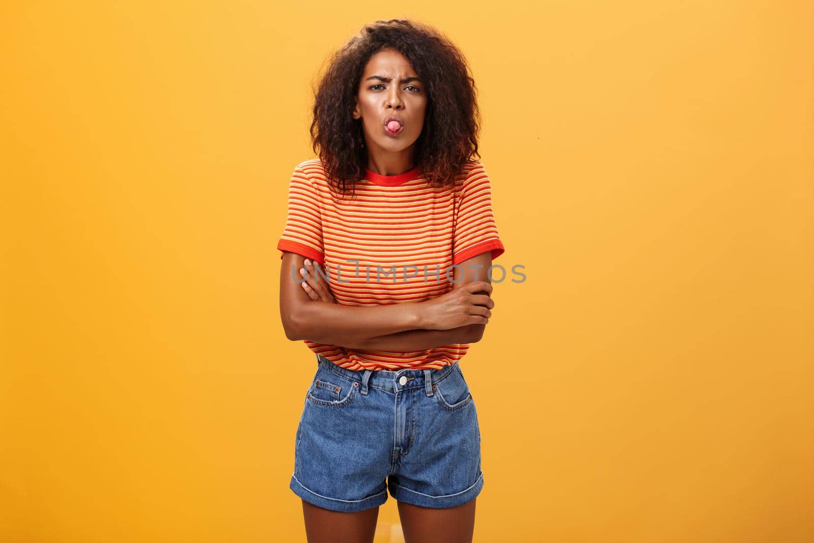 Immature girl showing bad side of character. Portrait of childish offended or displeased young African-American woman with curly hair showing tongue crossing arms on chest over orange background.