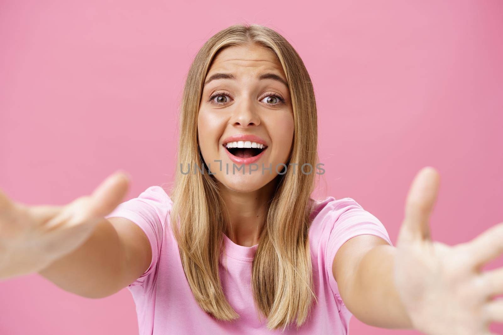 Close-up shot of caring and loving silly girl with tanned skin and fair hair pulling hands towards camera to give warm hug smiling broadly gazing with admiration wanting cuddle over pink background by Benzoix