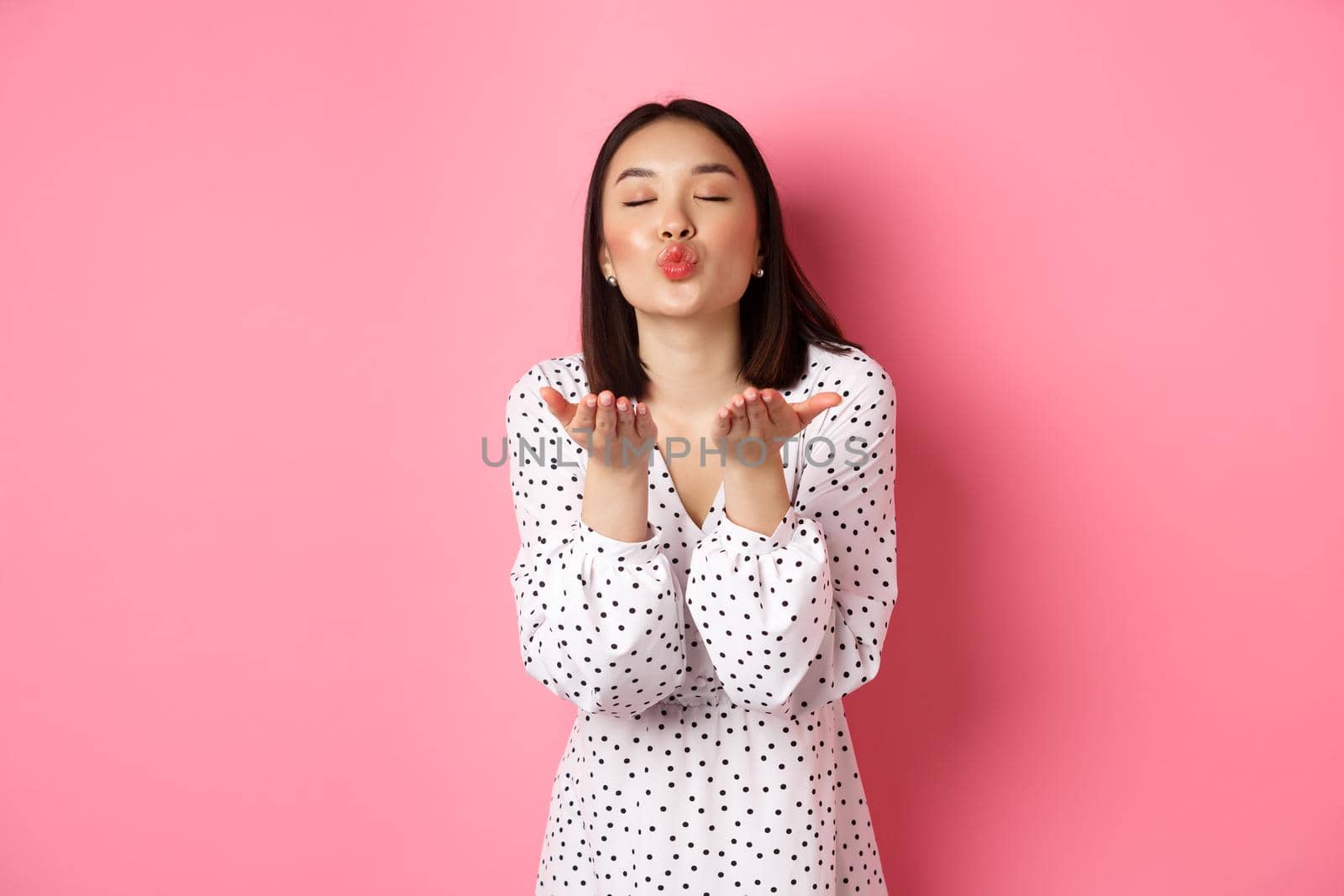 Beautiful chinese girl in dress blowing air kisses, sending mwah at camera, standing over pink background by Benzoix