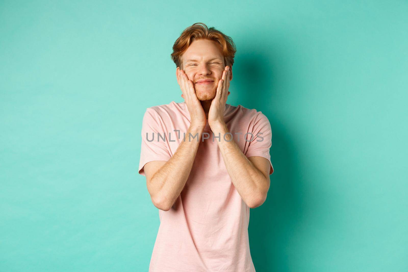 Image of cute and silly young man with red hair, feeling satisfaction while touching his face, smiling happy, standing over turquoise background by Benzoix