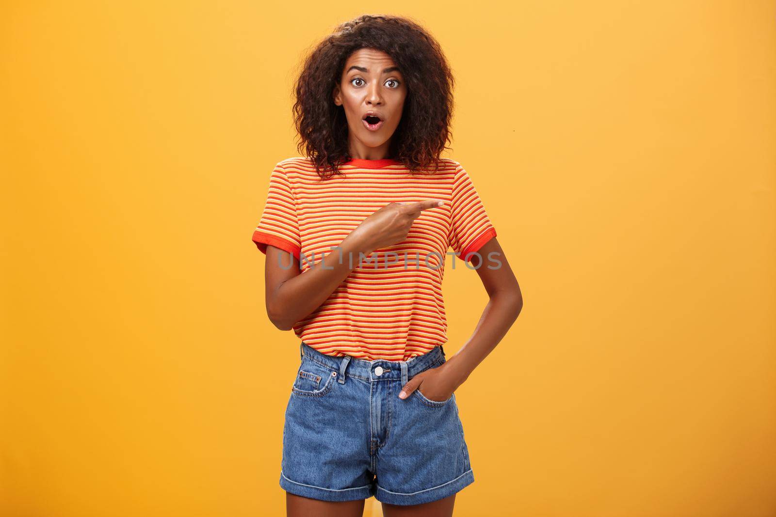 Studio shot of stylish fit female model in denim shorts and t-shirt gasping impressed pointing right while asking question being interested during observing masterclass over orange background. Lifestyle.