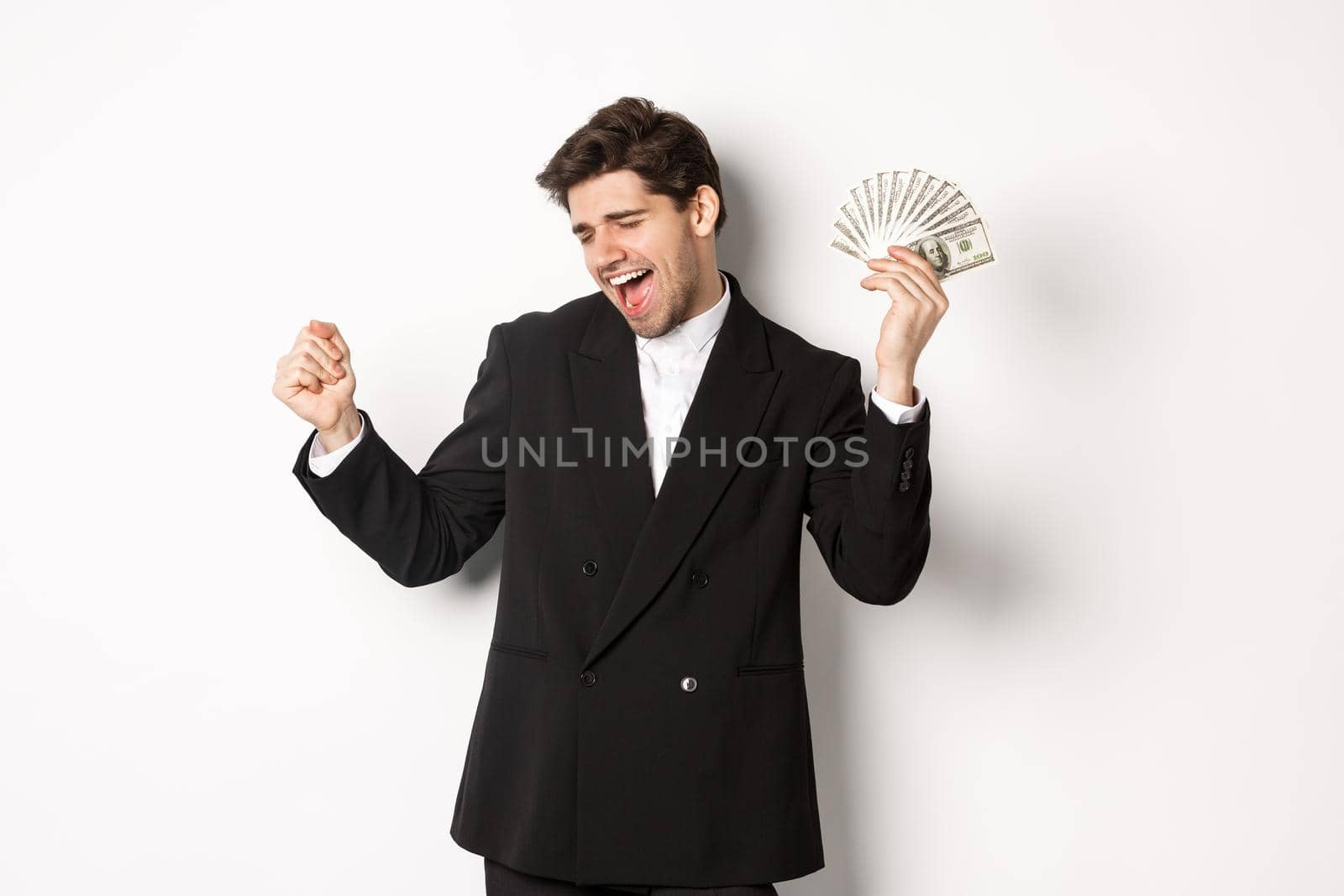 Portrait of handsome and successful businessman in suit, dancing with money, standing against white background.