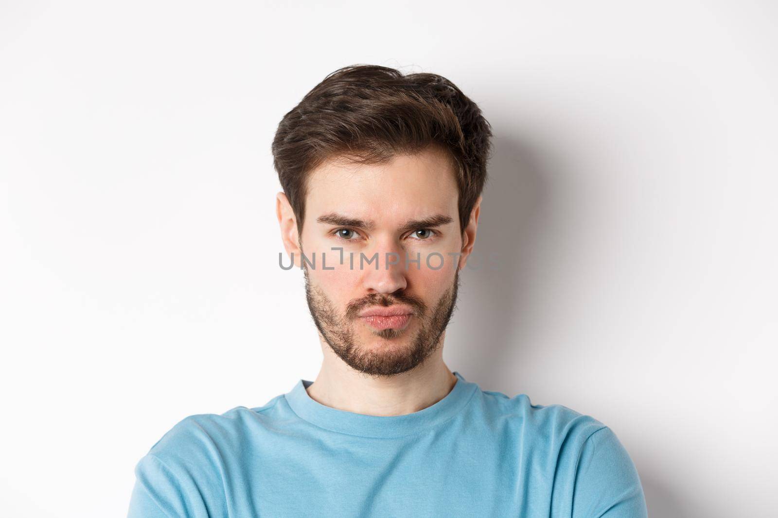 Close-up of grumpy young man grimacing, sulking and making offended face, standing upset over white background.