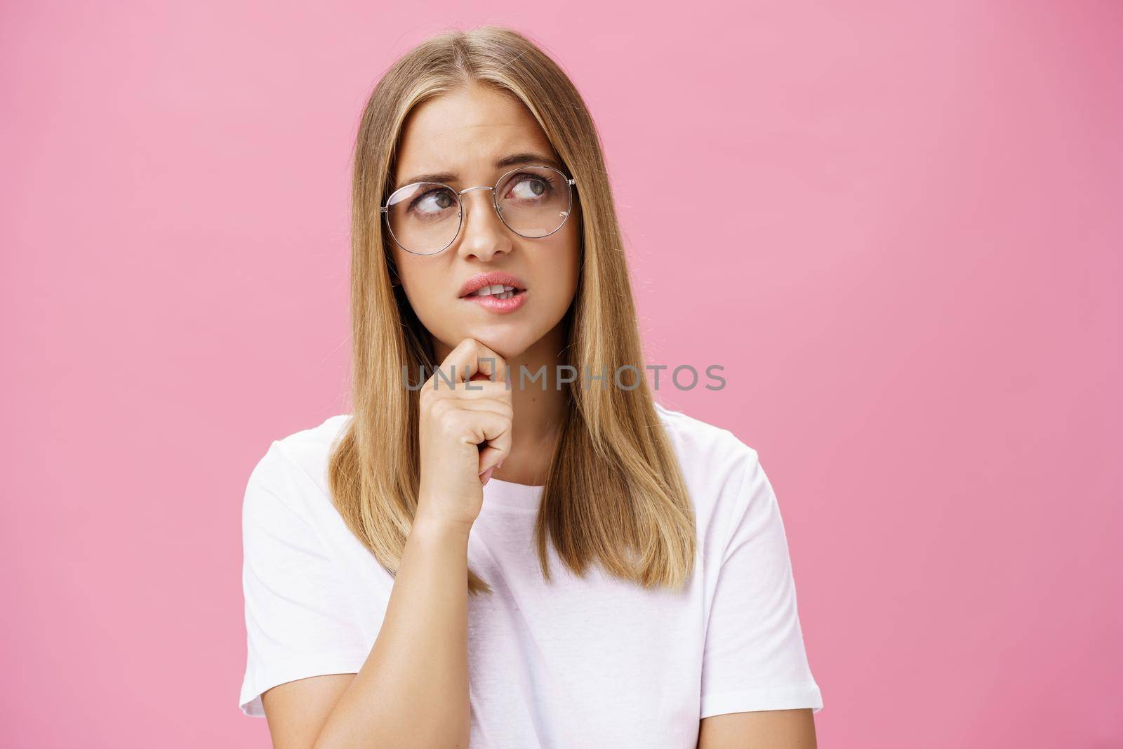 Studio shot of insecure smart nerdy woman in glasses and white t-shirt standing troubled and worried touching chin looking bothered at upper right corner hesitating, thinking against pink wall.