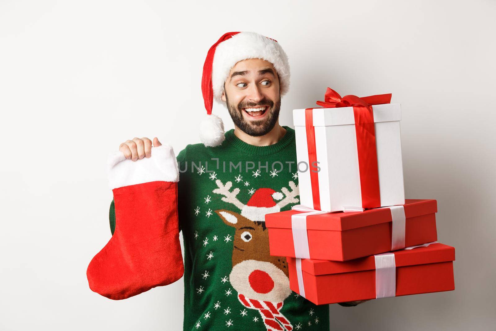 Xmas and winter holidays concept. Excited man holding christmas sock and gift boxes, celebrating New Year, standing over white background.