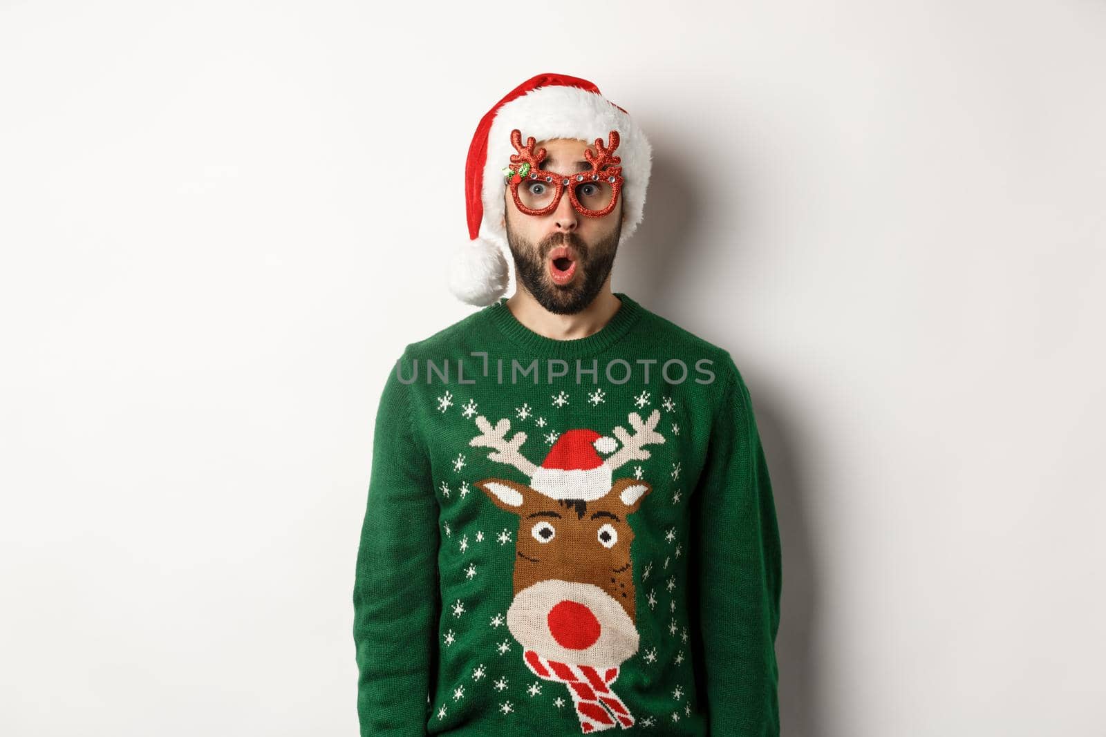 Christmas holidays, celebration concept. Surprised man in Santa hat and party glasses looking at something awesome, standing over white background.