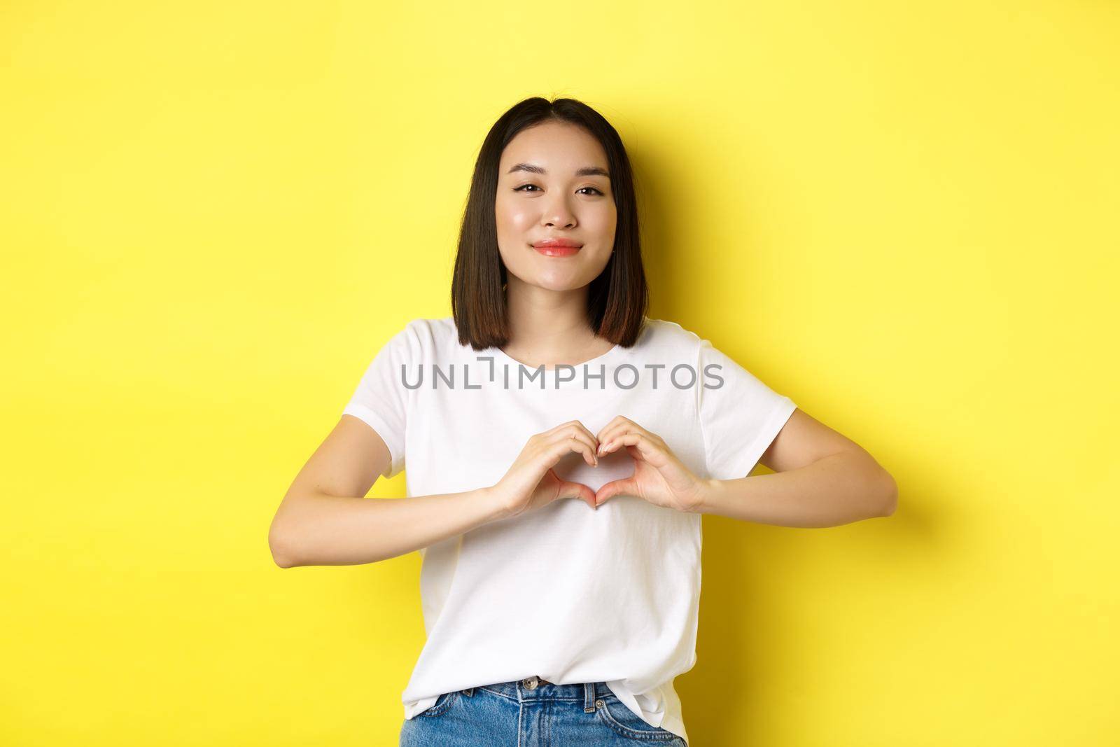 Beautiful asian woman showing I love you heart gesture, smiling at camera, standing against yellow background. Concept of valentines day and romance.