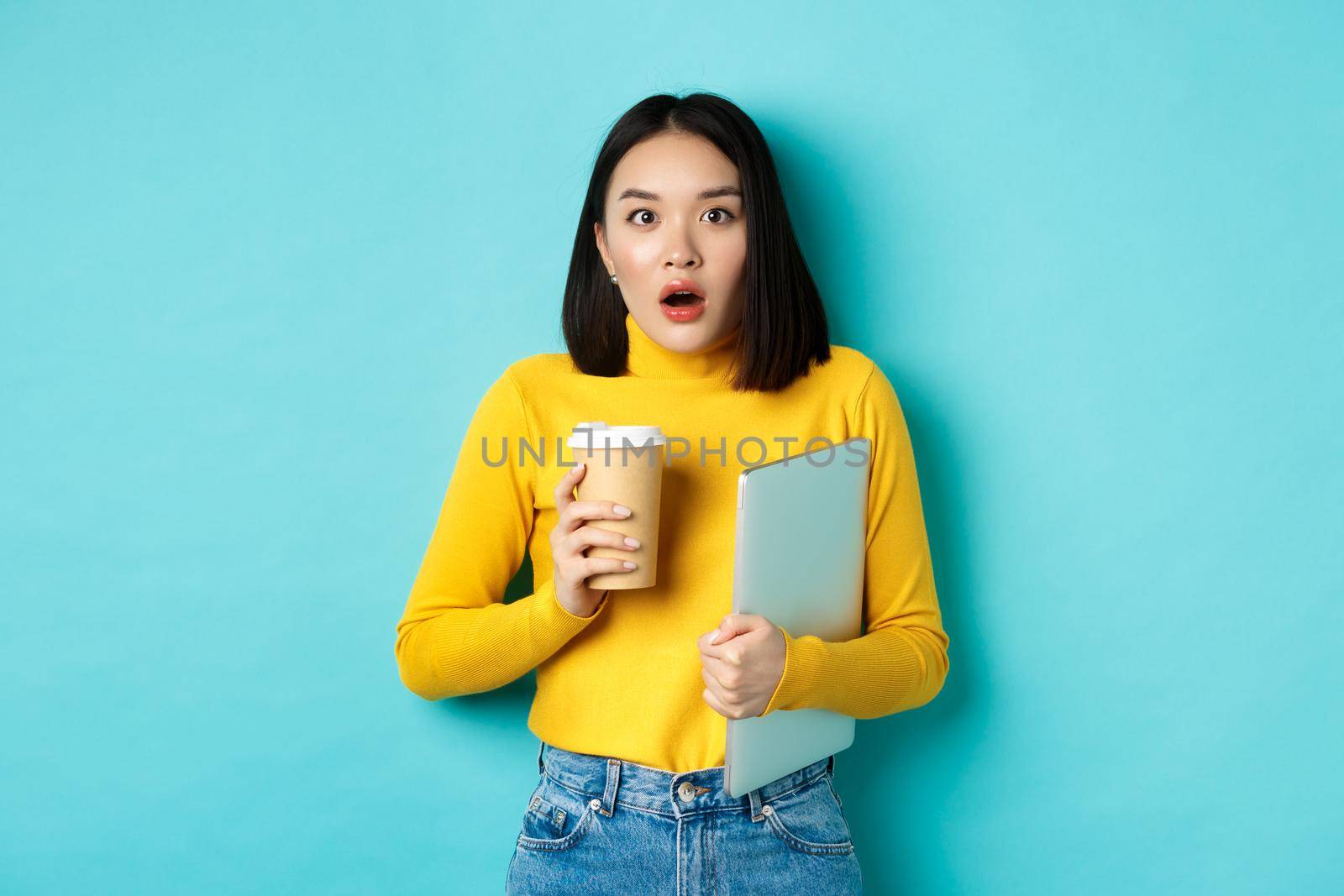 Shocked asian woman employee, drinking coffee from cafe takeaway, holding laptop, gasping and staring amazed at camera, standing over blue background.