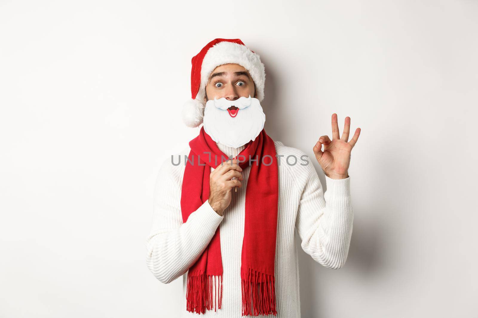 Christmas party and celebration concept. Happy male model in Santa Claus hat and white beard mask, showing ok gesture, standing over white background.