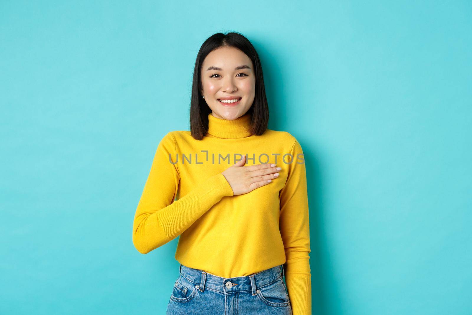Image of proud smiling asian woman holding hand on heart, showing respect to national anthem, standing over blue background by Benzoix