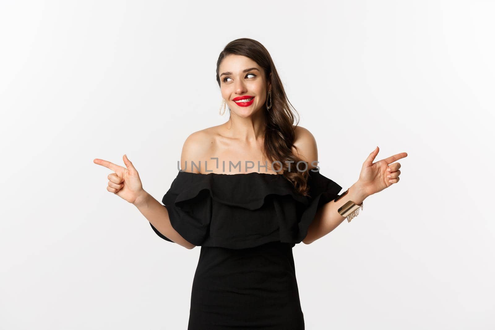 Fashion and beauty. Attractive woman in jewelry, makeup and black dress, smiling and pointing fingers sideways copy space offer, white background.