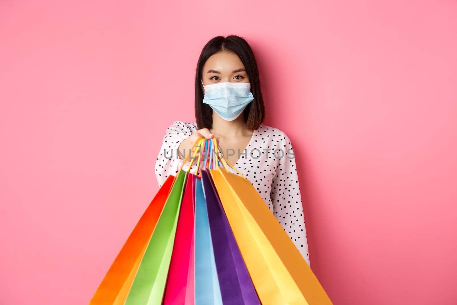 Covid-19, pandemic and lifestyle concept. Beautiful asian woman in face mask giving shopping bags, social distance in store, standing over pink background.