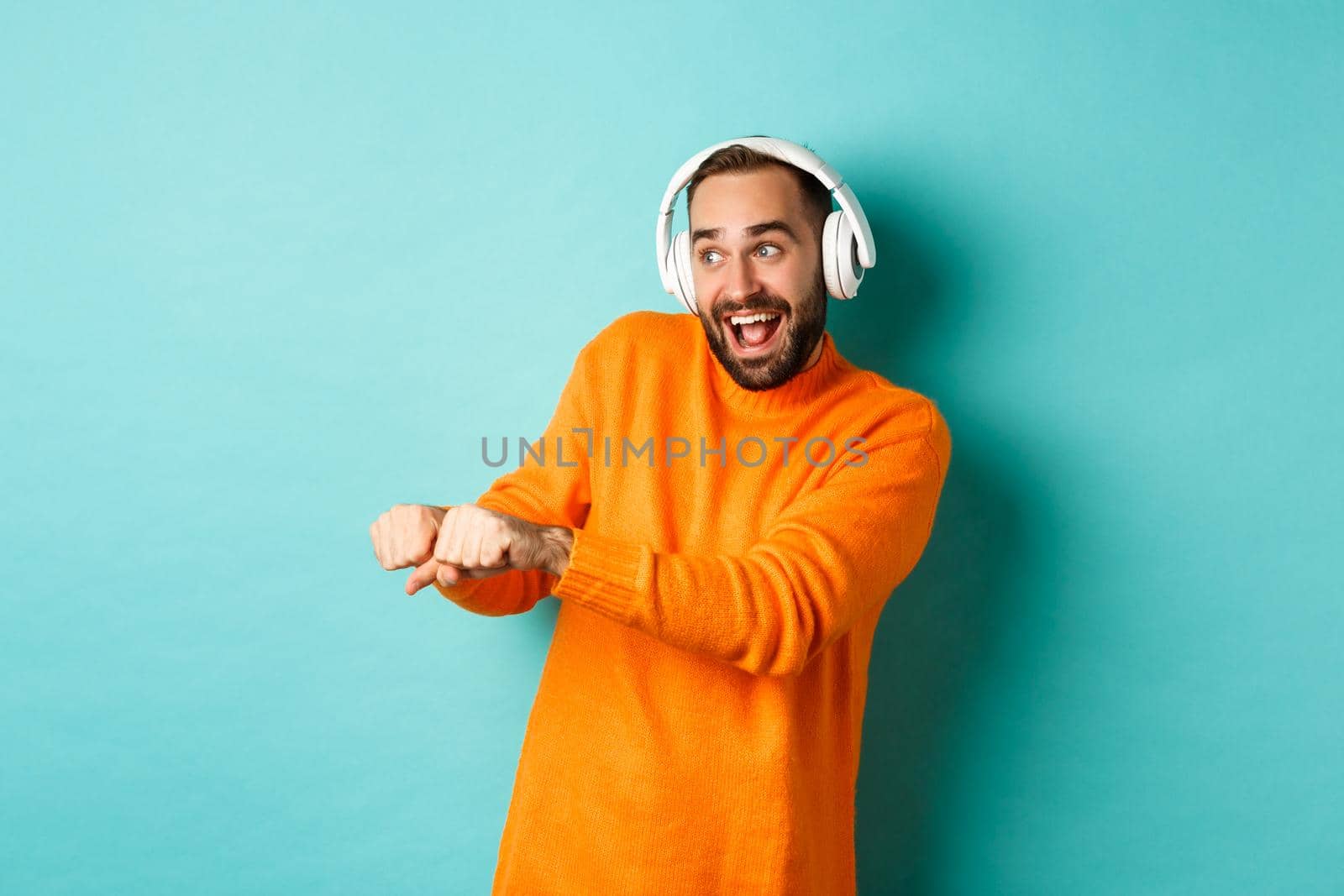 Happy man listening music in headphones and dancing funny, standing over turquoise background. Copy space