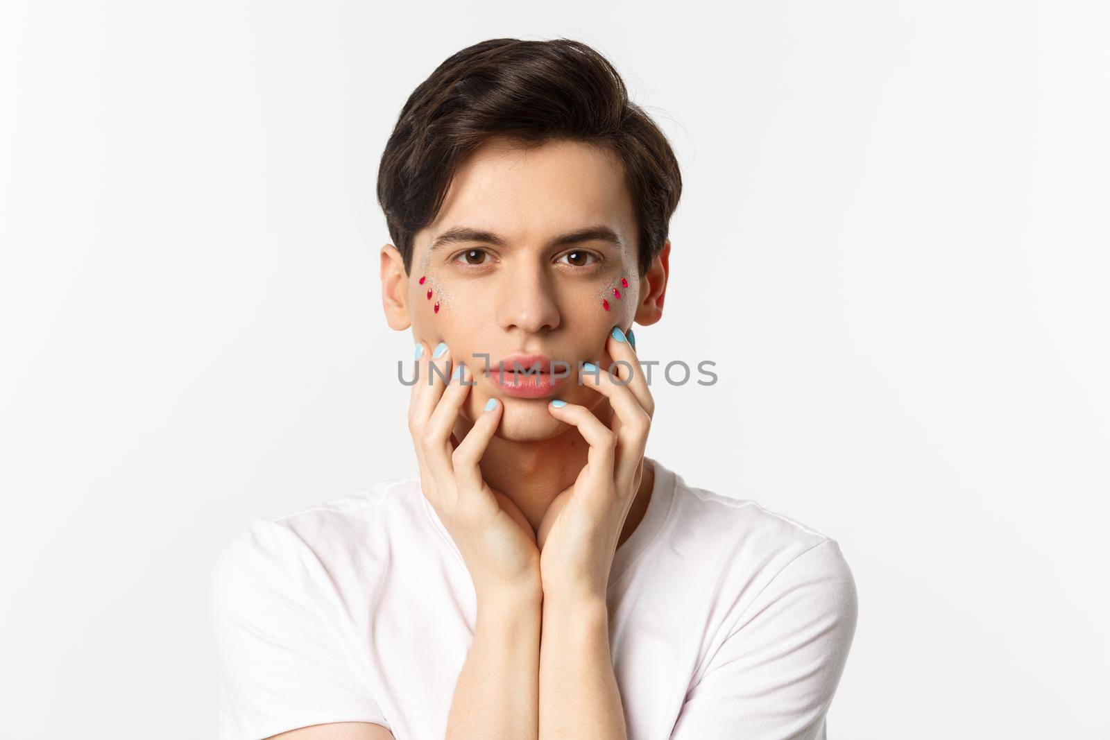 People, lgbtq and beauty concept. Close-up of beautiful queer man touching face with fingers with blue nail polish, standing over white background.