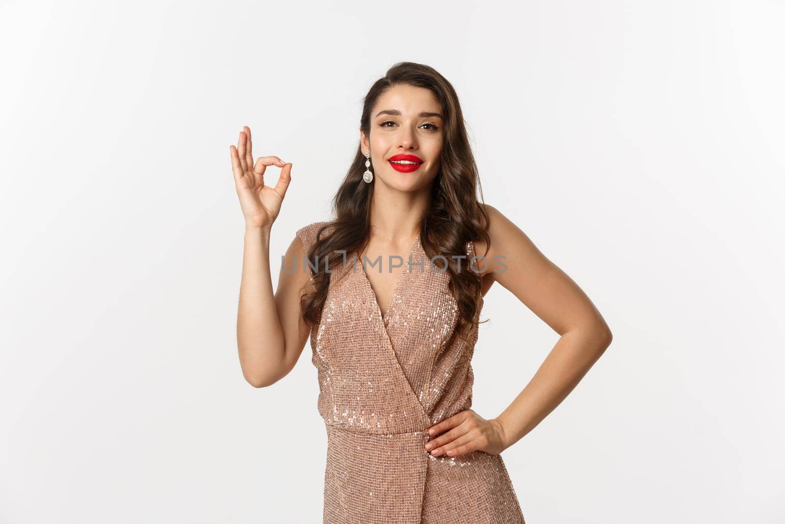 Beautiful young woman with red lipstick, wearing glamour glitter dress showing okay sign, approve and like product, praise good work, standing over white background.