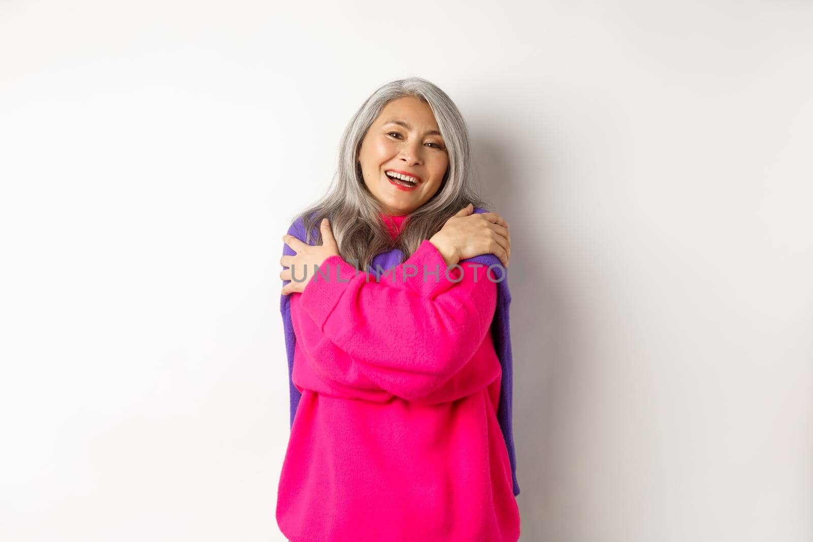 Valentines day and holidays concept. Lovely asian senior woman in pink sweater hugging herself with eyes closed, smiling, standing over white background.