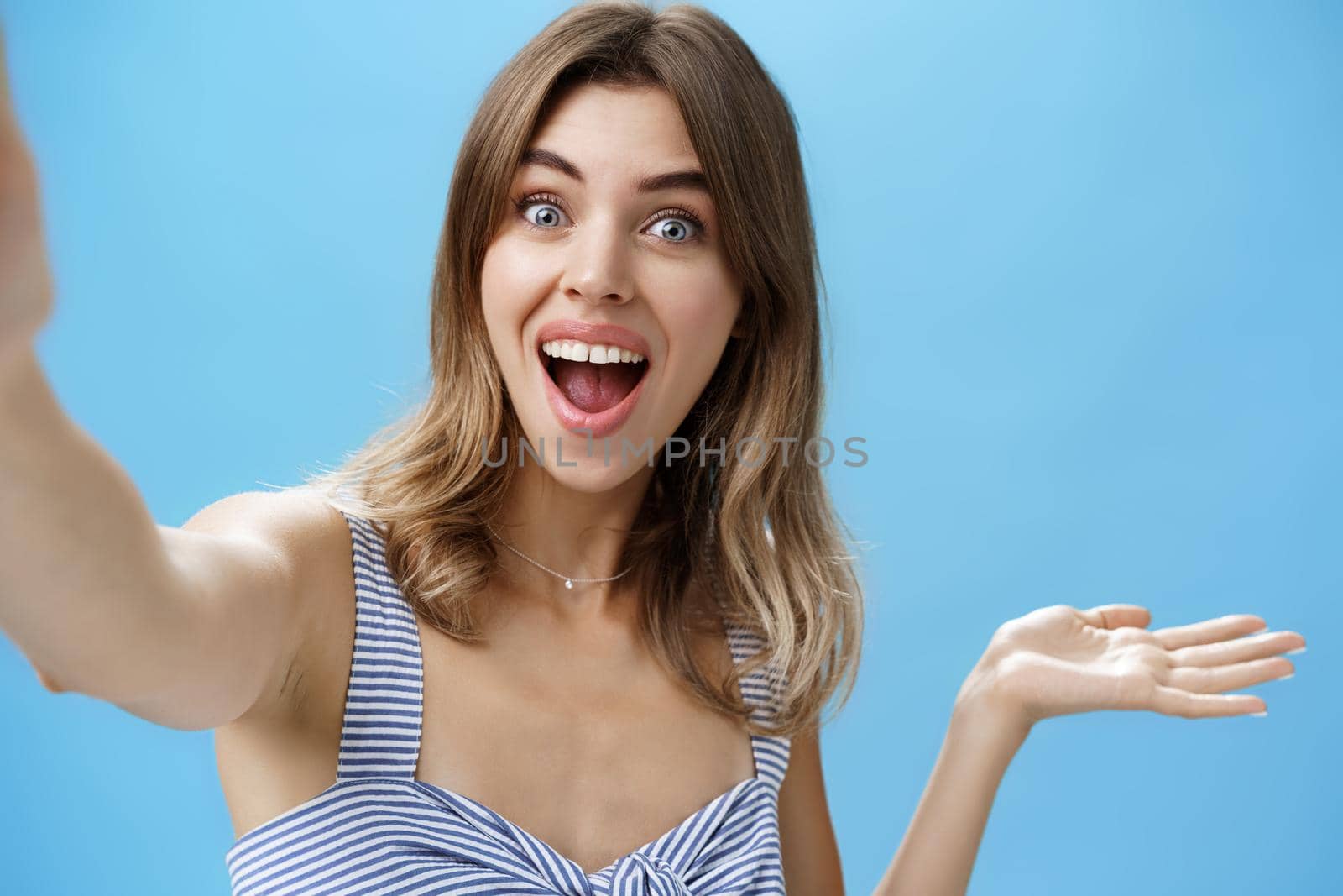 Waist-up shot of charismatic upbeat enthusiastic cute female blogger raising hand over copy space and holding camera as if recording video from vacation showing cool product over blue background. Lifestyle.