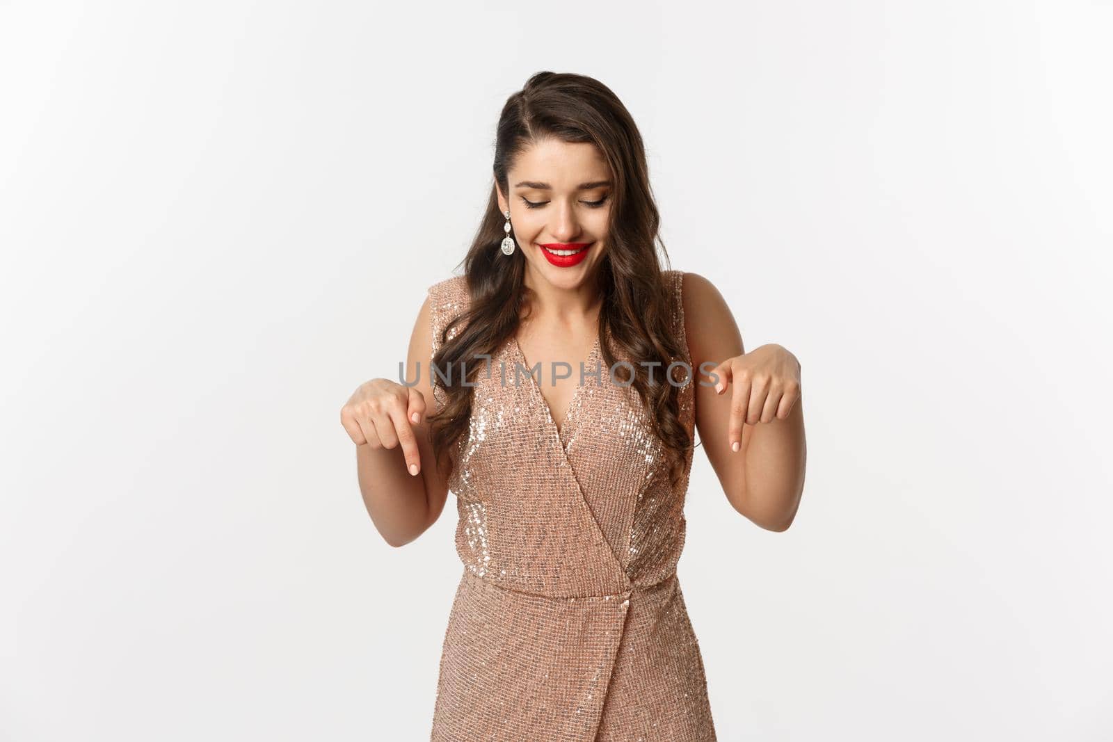 Beautiful young woman with red lipstick, wearing elegant dress for christmas party, pointing fingers down and smiling pleased, standing over white background by Benzoix