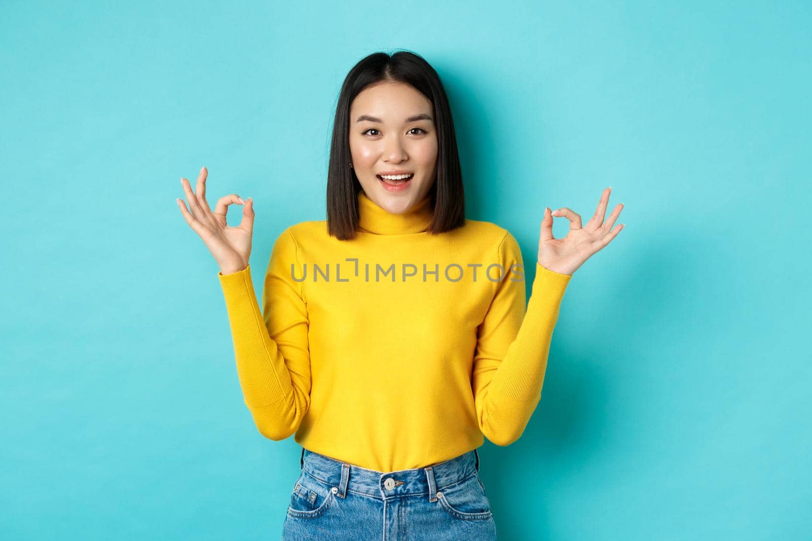 Cheerful asian female model showing okay gestures, smiling and looking impressed, praise product, standing over blue background.