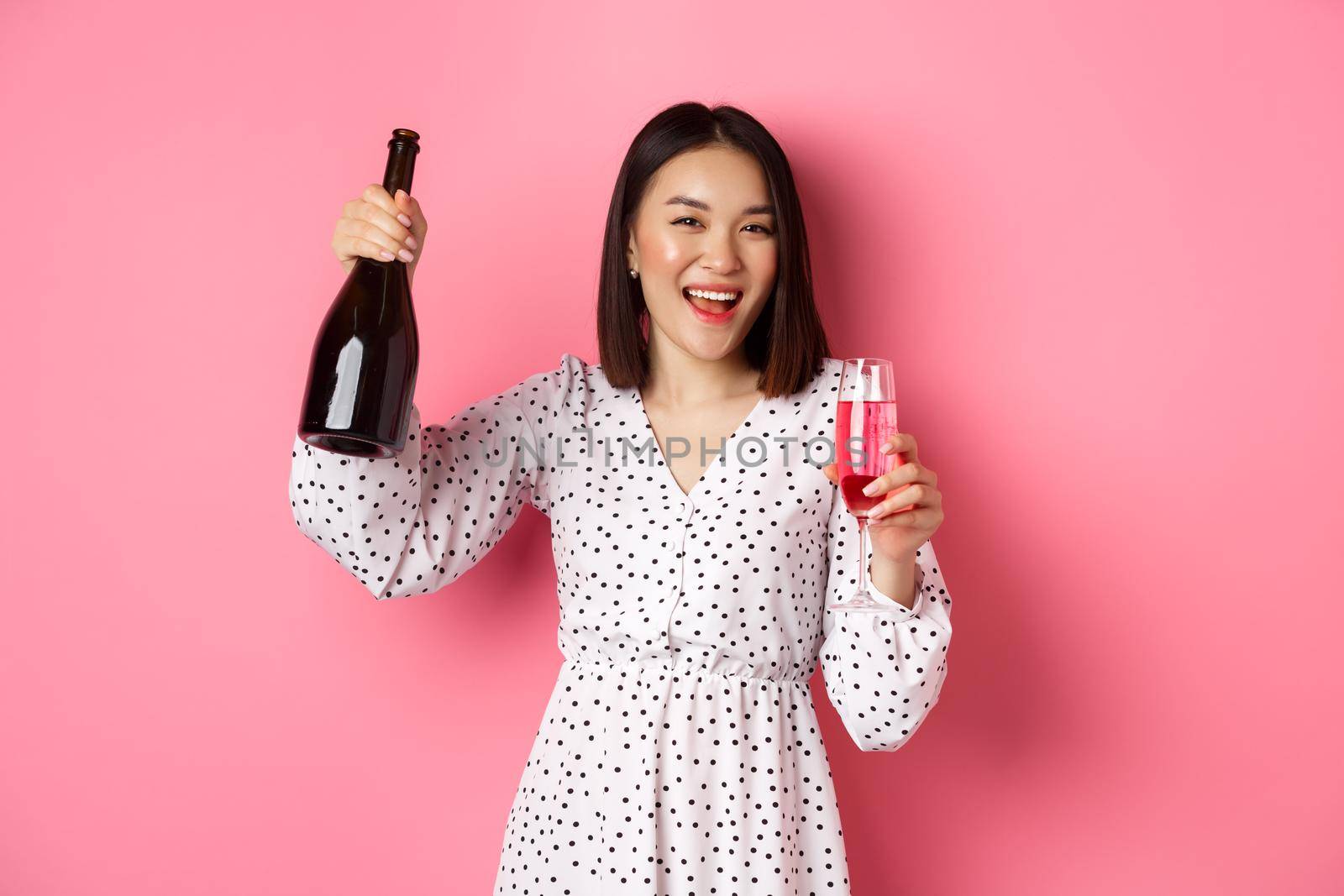 Happy asian woman celebrating, having fun and partying, pouring glass of champagne and laughing, standing over pink background.