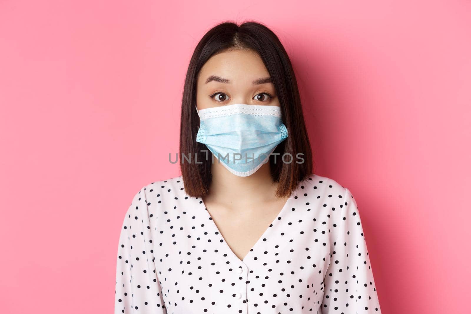 Covid-19, pandemic and lifestyle concept. Surprised asian woman in face mask raising eyebrows, staring at camera amazed, standing against pink background by Benzoix