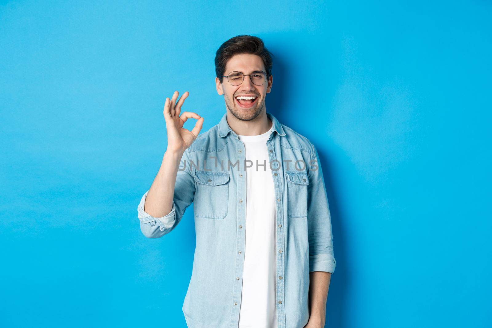 Confident smiling man in glasses showing ok sign, winking to guarantee or recommend something, blue background.