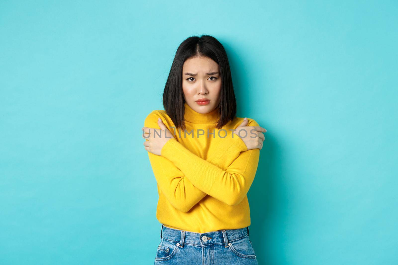 Young asian woman feeling like victim, cover body with hands and looking anxious at camera, standing over blue background.