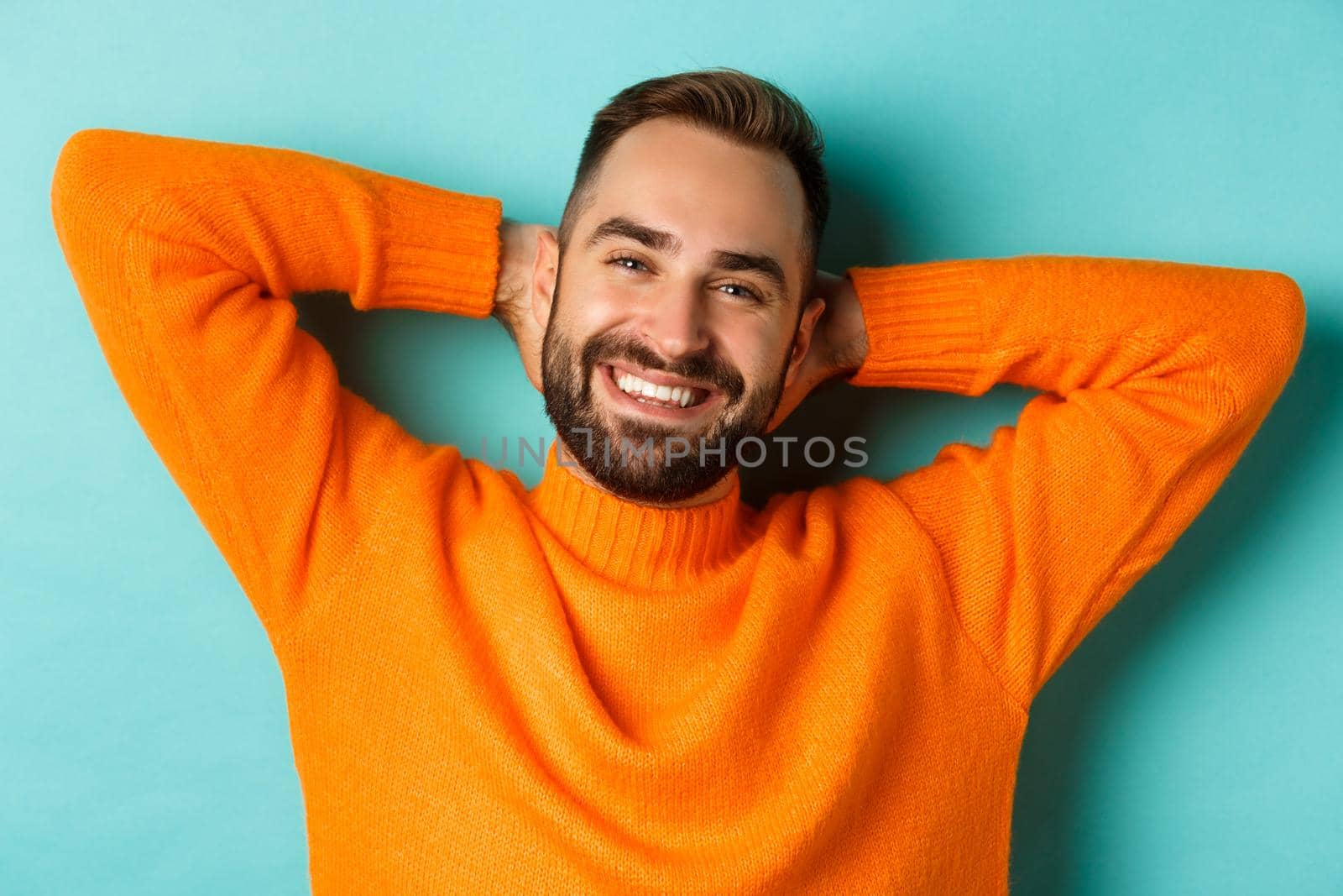 Carefree young man daydreaming, resting with hands behind head and looking camera happy, standing over turquoise background in orange sweater.