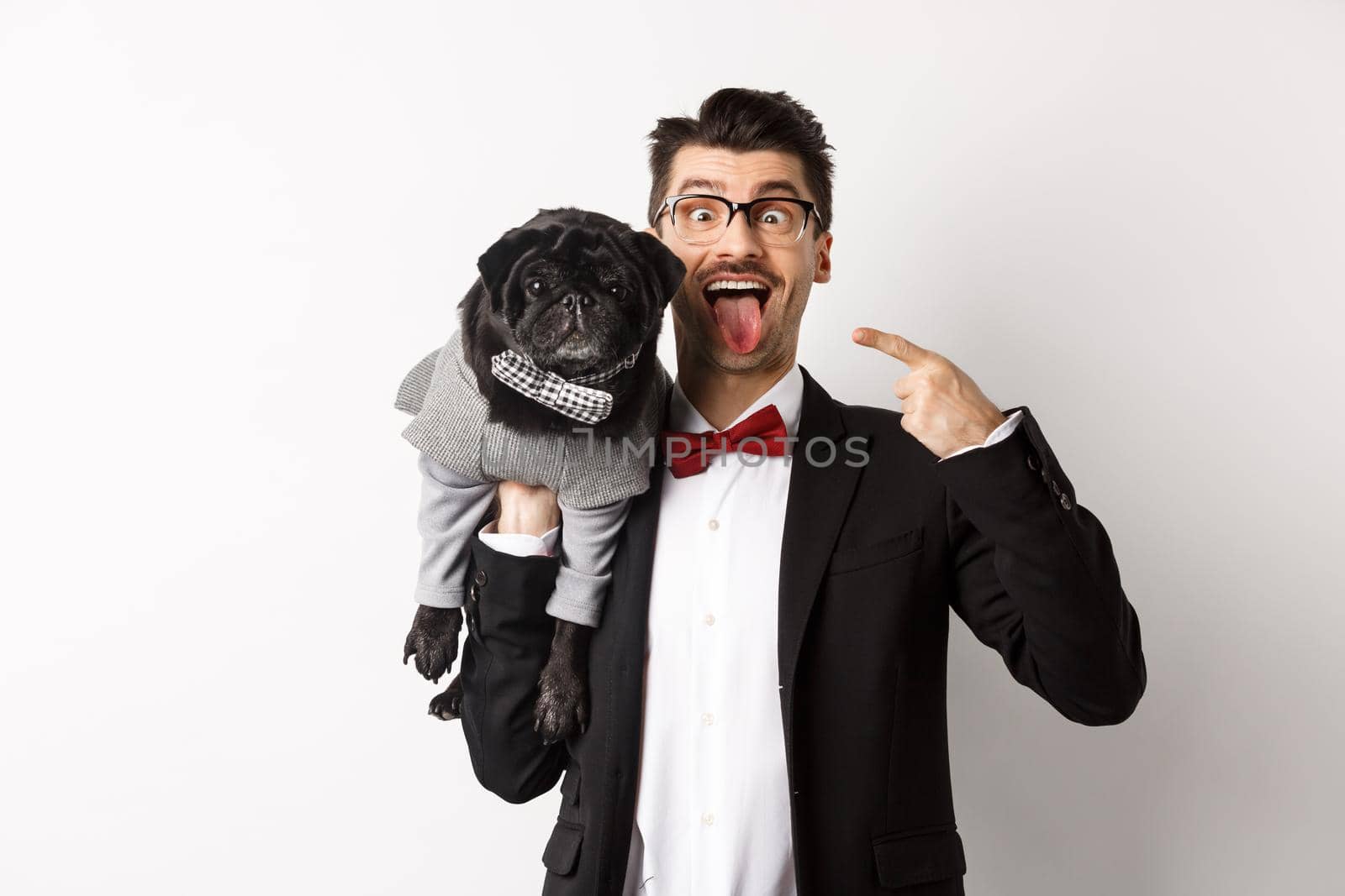 Funny young man in party suit, showing tongue and making grimaces, pointing at cute black dog in winter clothes, standing over white background.
