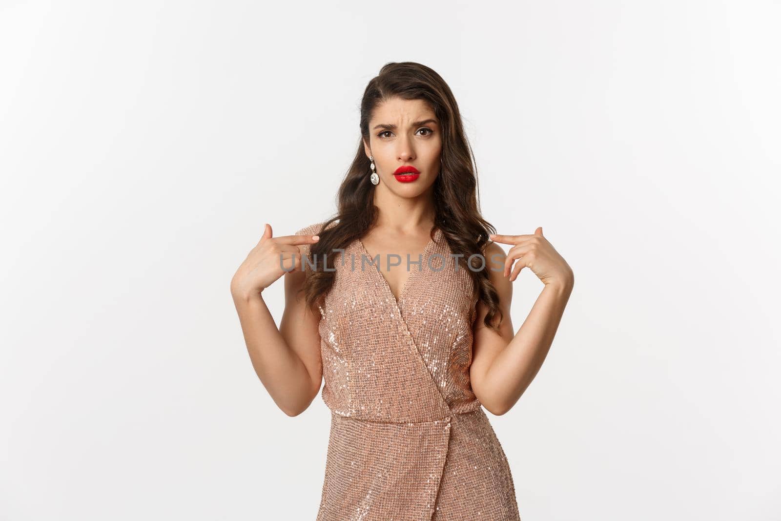 Self-assured woman looking confused and displeased while pointing at herself, standing in glamour dress for new year party, white background.