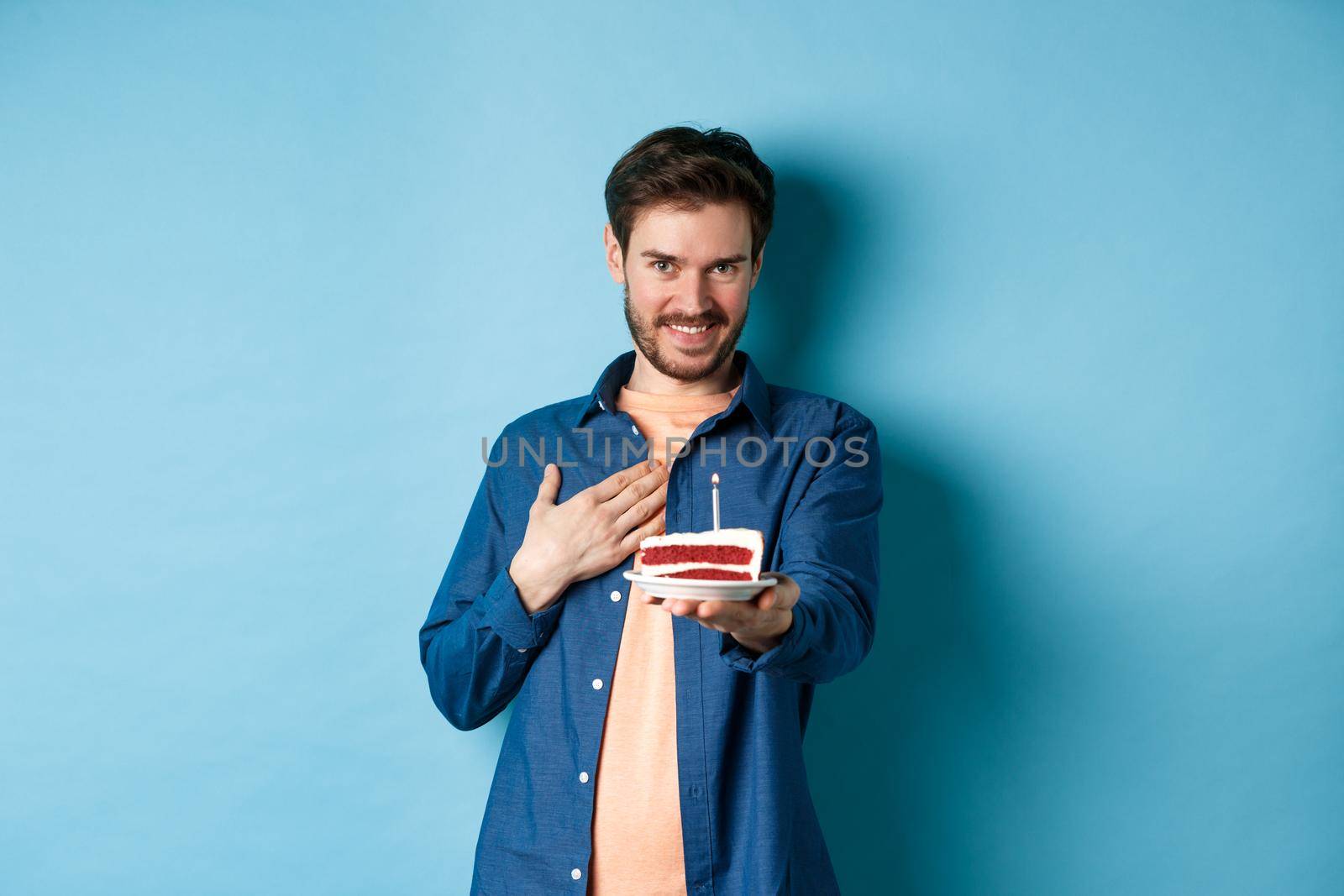 Celebration and holiday concept. Man blushing and wishing happy birthday, stretch out b-day cake with lit candle, standing on blue background by Benzoix