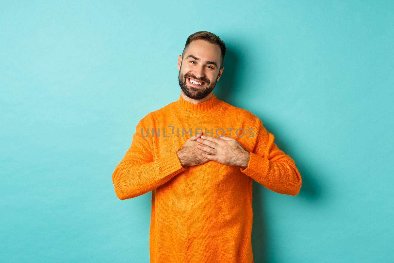 Grateful man smiling, holding hands on heart, thank you gesture, feeling touched with gift, standing over turquoise background.