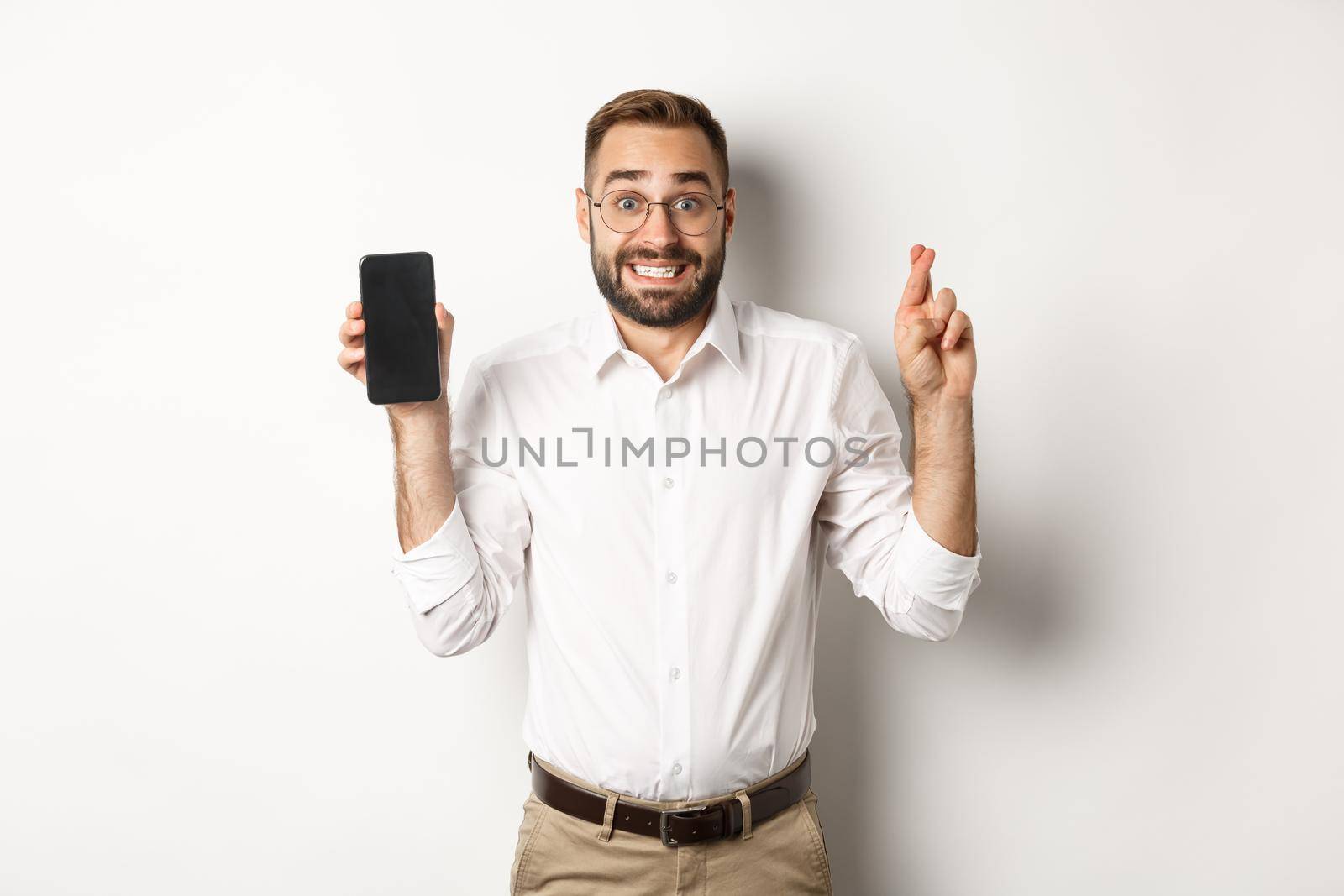 Hopeful young business man showing mobile screen, holding fingers crossed, waiting for online results, standing over white background.