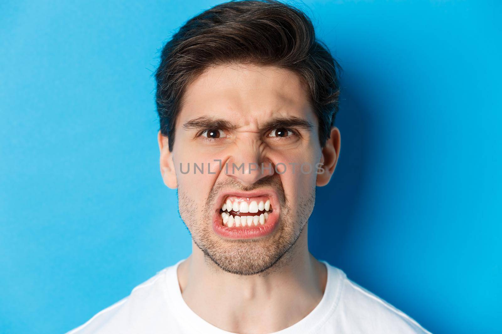 Headshot of angry caucasian man looking with scorn and dismay, being mad at person, standing against blue background.