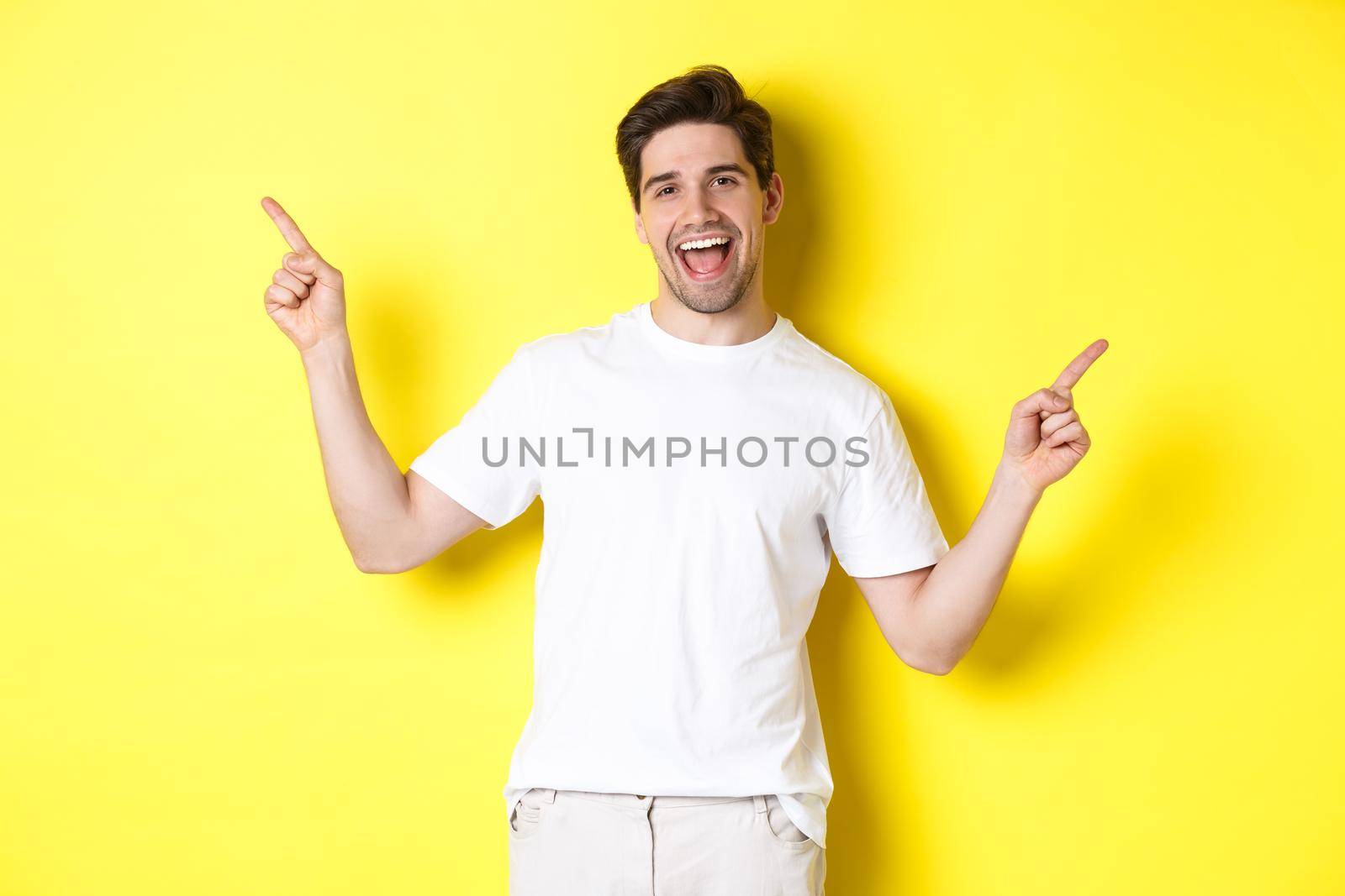 Happy stylish guy showing two variants, pointing fingers sideways at left and right promo, standing over yellow background.