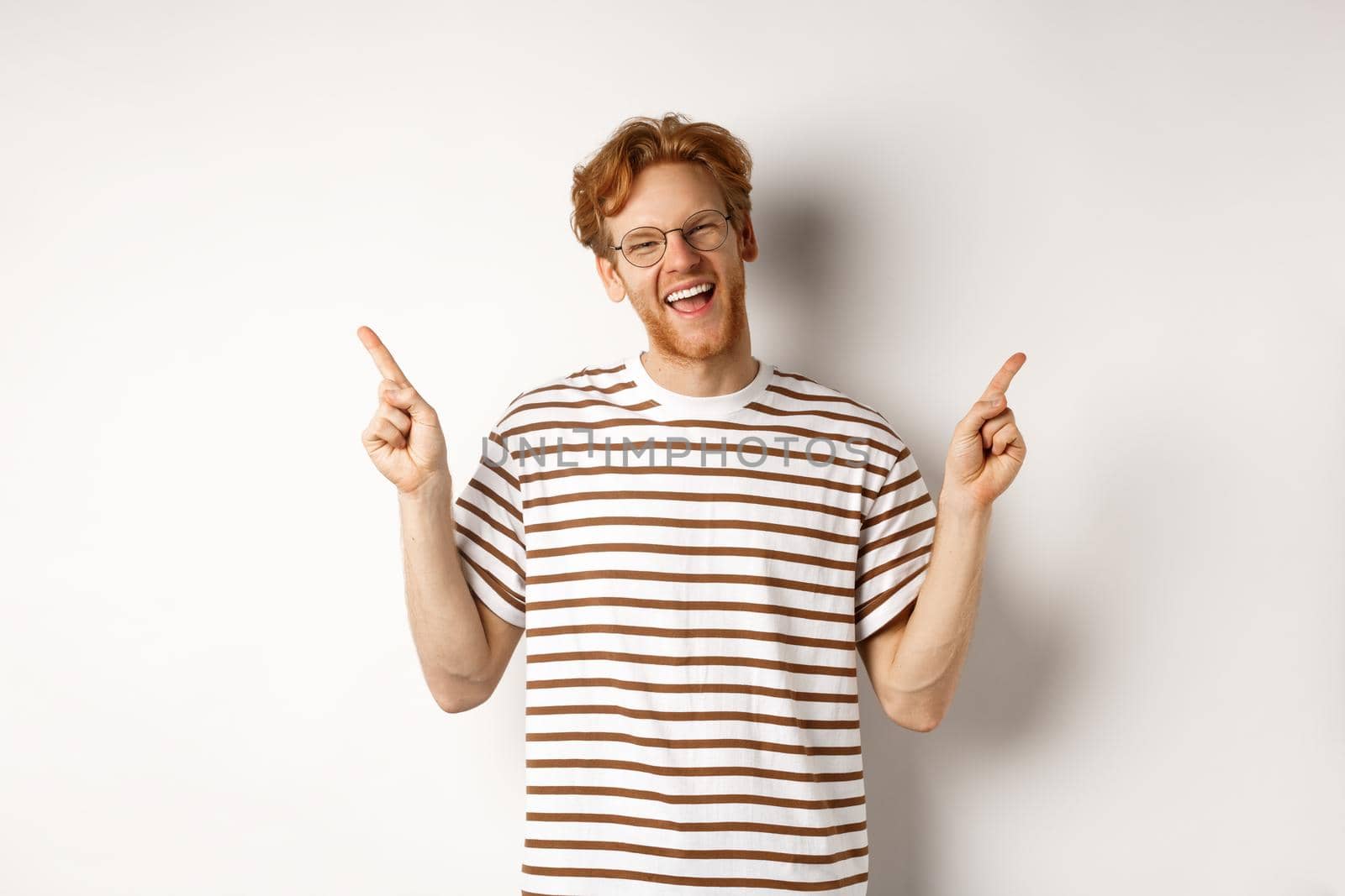 Happy hipster guy with red messy hair and glasses smiling, pointing fingers up, showing sale promotion, white background.