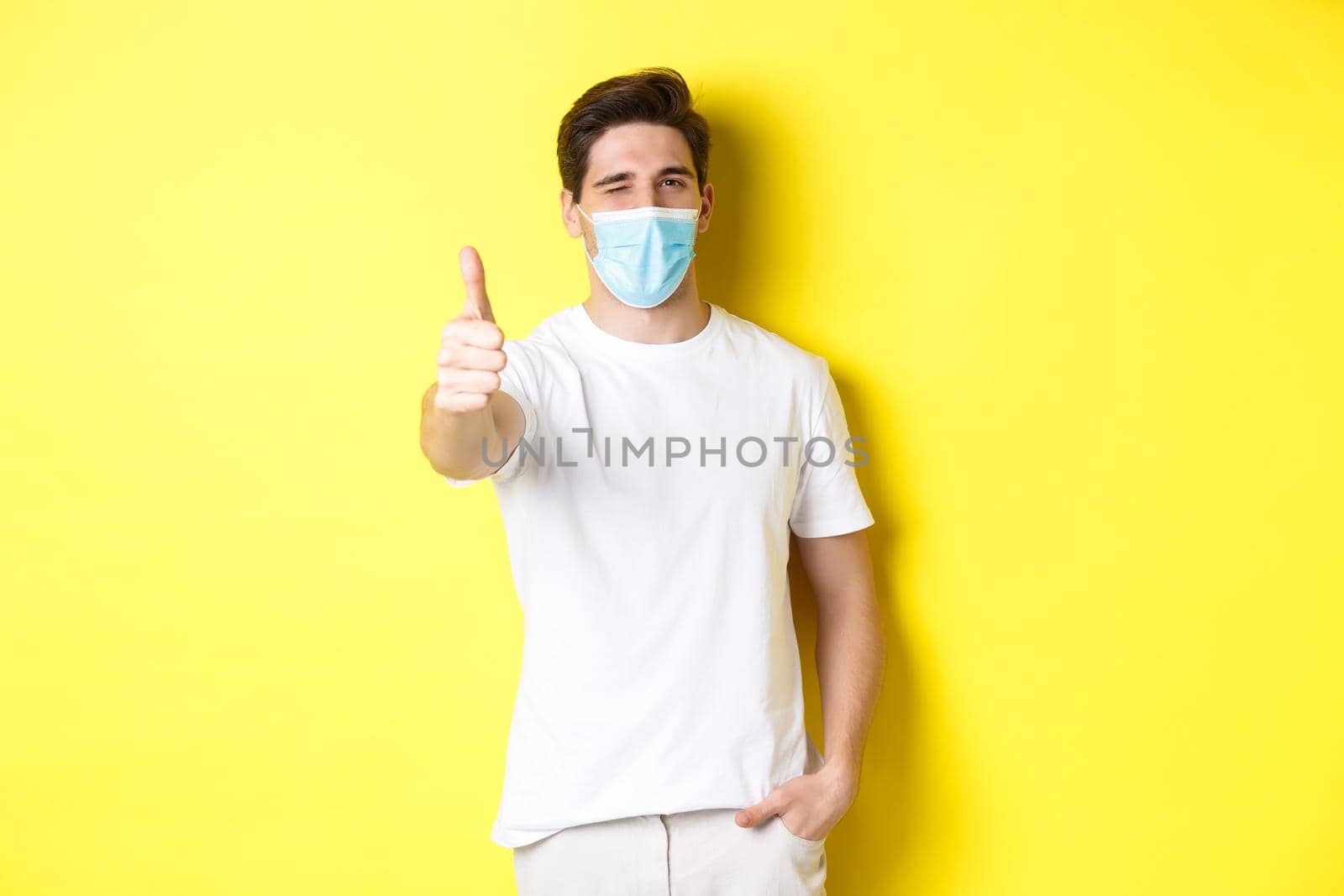 Concept of coronavirus, pandemic and social distancing. Confident young man in medical mask showing thumbs up and winking, yellow background by Benzoix