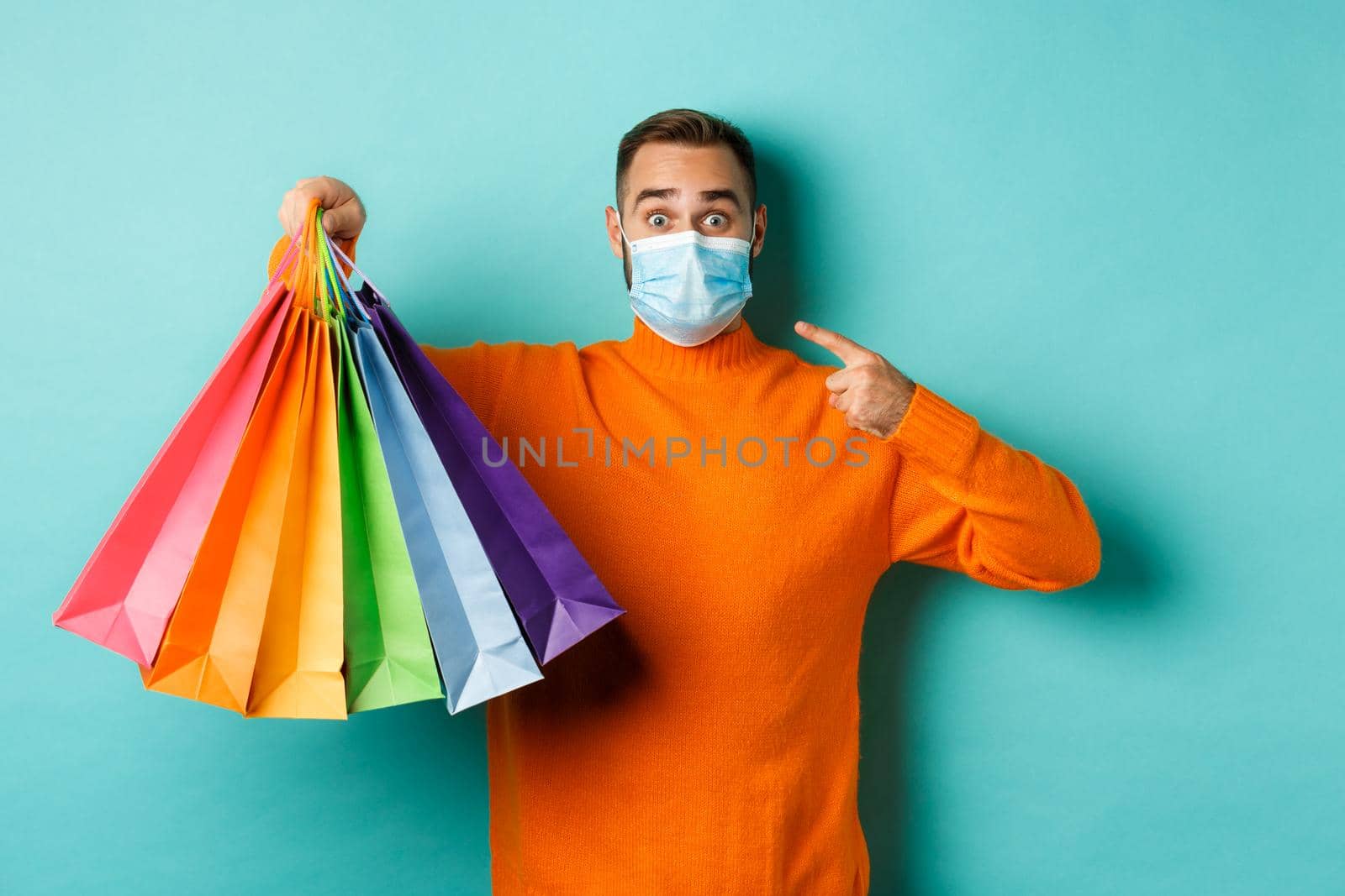 Covid-19, social distancing and lifestyle concept. Young man in face mask showing shopping bags, buying holiday gifts during pandemic, standing over blue background.